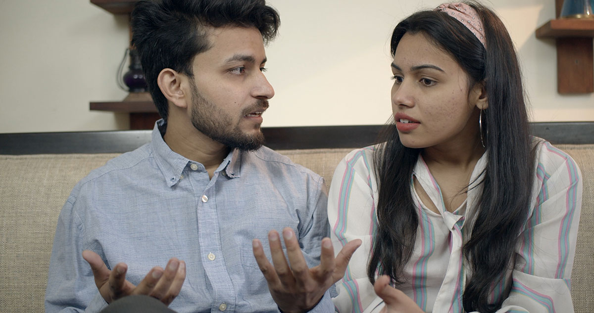 A young Indian couple sitting on a sofa, talking, and looking serious. He is spreading his hands out and she is looking puzzled.
